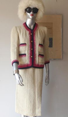 Vintage Chanel-style cream boucle skirt suit featuring pink and navy trim with gold-tone buttons, displayed on a silver mannequin.