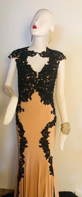 Elegant floor-length evening gown in a nude or light tan fabric, featuring intricate black lace applique detailing along the bodice,...