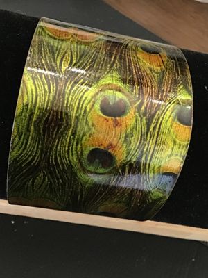Sale Photo Thumbnail #37: A wide metal cuff bracelet featuring a vivid glossy peacock feather pattern on the exterior with a gold-tone brass interior finish. Measures approximately 1.75 to 2 inches wide. Shows minor wear along the edges and interior surface, but overall in good, w