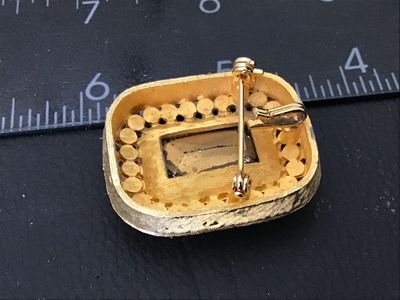 Sale Photo Thumbnail #70: Vintage gold-tone brooch featuring a large, emerald-cut red glass/rhinestone center stone surrounded by a halo of clear pave rhinestones. Textured brushed gold-tone finish with pin back mechanism and hook attachment. Measures approximately 1.5 inches wide