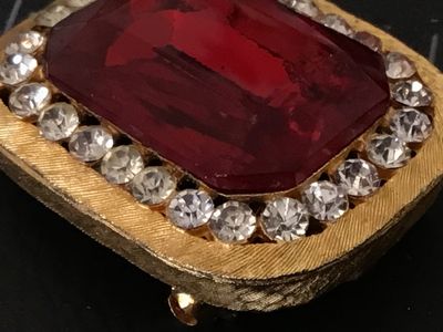 Sale Photo Thumbnail #72: Vintage gold-tone brooch featuring a large, emerald-cut red glass/rhinestone center stone surrounded by a halo of clear pave rhinestones. Textured brushed gold-tone finish with pin back mechanism and hook attachment. Measures approximately 1.5 inches wide
