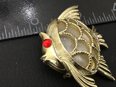 Sale Photo Thumbnail #74: A vintage gold-tone textured fish brooch featuring a red rhinestone eye and a central faux pearl or lucite cabochon inset. Measures approximately 1.5 inches wide. Shows minor age-appropriate surface wear; pin back and clasp are intact and functional.