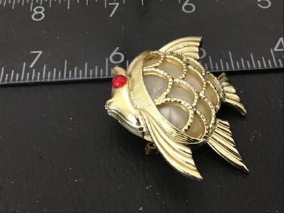 Sale Photo Thumbnail #76: A vintage gold-tone textured fish brooch featuring a red rhinestone eye and a central faux pearl or lucite cabochon inset. Measures approximately 1.5 inches wide. Shows minor age-appropriate surface wear; pin back and clasp are intact and functional.