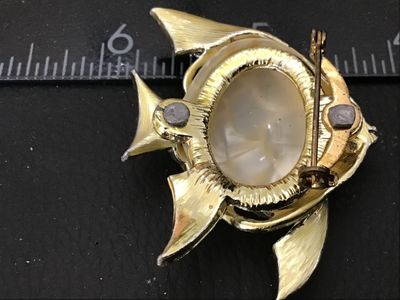Sale Photo Thumbnail #75: A vintage gold-tone textured fish brooch featuring a red rhinestone eye and a central faux pearl or lucite cabochon inset. Measures approximately 1.5 inches wide. Shows minor age-appropriate surface wear; pin back and clasp are intact and functional.