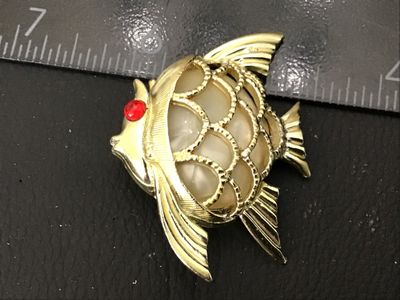 Sale Photo Thumbnail #73: A vintage gold-tone textured fish brooch featuring a red rhinestone eye and a central faux pearl or lucite cabochon inset. Measures approximately 1.5 inches wide. Shows minor age-appropriate surface wear; pin back and clasp are intact and functional.
