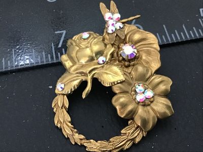Sale Photo Thumbnail #81: A vintage gold-tone metal brooch featuring a wreath design adorned with sculpted flowers, leaves, and a dragonfly detail. Accentuated with aurora borealis (AB) iridescence rhinestones. Standard pin-back closure in working order. Measures approximately 2 i