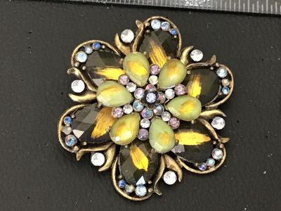 Sale Photo Thumbnail #89: Ornate vintage floral-form brooch featuring layered green and smoked glass stones with gold-tone foil details, accented by iridescent aurora borealis and lavender rhinestones. Set in an antiqued brass-tone metal setting. Includes a standard roll-over safe