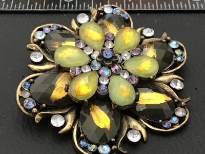 Sale Photo Thumbnail #88: Ornate vintage floral-form brooch featuring layered green and smoked glass stones with gold-tone foil details, accented by iridescent aurora borealis and lavender rhinestones. Set in an antiqued brass-tone metal setting. Includes a standard roll-over safe