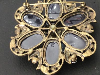 Sale Photo Thumbnail #90: Ornate vintage floral-form brooch featuring layered green and smoked glass stones with gold-tone foil details, accented by iridescent aurora borealis and lavender rhinestones. Set in an antiqued brass-tone metal setting. Includes a standard roll-over safe