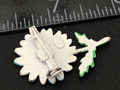 Sale Photo Thumbnail #92: A vintage 1960s-1970s style enamel flower brooch featuring a light blue and white daisy with a yellow center, green stem, and leaves. Made of painted metal with a standard pinback closure. Shows some wear with light enamel chipping on the petals and stem.