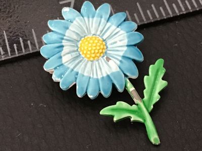 Sale Photo Thumbnail #91: A vintage 1960s-1970s style enamel flower brooch featuring a light blue and white daisy with a yellow center, green stem, and leaves. Made of painted metal with a standard pinback closure. Shows some wear with light enamel chipping on the petals and stem.