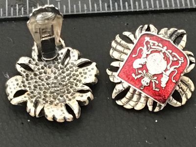 Sale Photo Thumbnail #100: Pair of vintage clip-on earrings featuring a textured silver-tone crown motif surrounding a diamond-shaped red enamel panel. The central shield showcases a detailed coat of arms/heraldic crest design. Silver-tone textured backs with functional clip closur