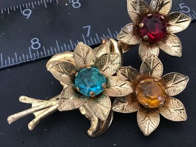 Sale Photo Thumbnail #117: Vintage gold-tone brooch featuring a floral bouquet design set with colorful round rhinestones in red, aqua blue, and amber yellow. The back features a standard pin bar closure. Measures approximately 3 inches in length. Shows vintage wear including patin