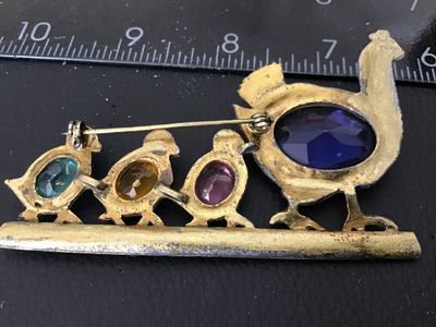 Sale Photo Thumbnail #147: A vintage gold-tone costume jewelry brooch depicting a mother hen with three chicks walking along a green painted bar. Features colorful faceted oval glass rhinestones in purple, pink, yellow, and blue, with tiny rhinestone eye details. Pin back clasp on 