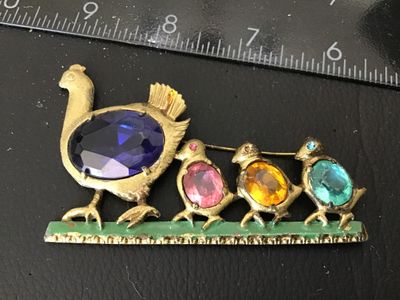 Sale Photo Thumbnail #144: A vintage gold-tone costume jewelry brooch depicting a mother hen with three chicks walking along a green painted bar. Features colorful faceted oval glass rhinestones in purple, pink, yellow, and blue, with tiny rhinestone eye details. Pin back clasp on 