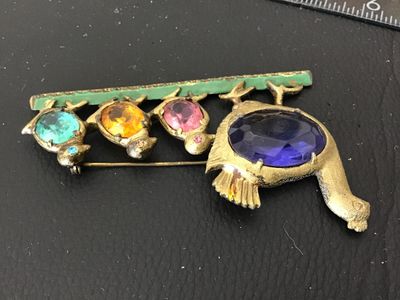 Sale Photo Thumbnail #146: A vintage gold-tone costume jewelry brooch depicting a mother hen with three chicks walking along a green painted bar. Features colorful faceted oval glass rhinestones in purple, pink, yellow, and blue, with tiny rhinestone eye details. Pin back clasp on 