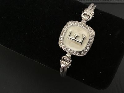 Sale Photo Thumbnail #23: Silver-tone twisted cable style cuff/bangle bracelet featuring a square center emblem with the initial 'E' set against an off-white enamel background, surrounded by a halo of clear rhinestones/crystals. Features a magnetic closure at the back. Light overa