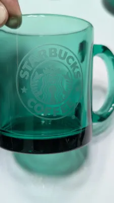 Sale Photo Thumbnail #285: This authentic vintage 1990s Starbucks mug is made in the USA from emerald-tinted, clear glass featuring an etched or embossed classic Siren logo