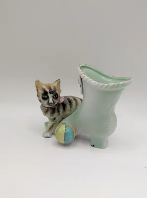 Sale Photo Thumbnail #352: The "UCAGCO China Handpainted Japan Cat, Boot, and Ball" is a charming mid-century vintage figurine. Made in Japan by the United China and Glass Company, it usually depicts a small, hand-painted ceramic kitten (often a tabby) either sitting on a boot/shoe