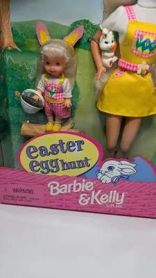 Sale Photo Thumbnail #366: Barbie Doll: Wears a vibrant yellow jumper with an Easter egg pattern pocket, a white t-shirt, a matching pink plaid hat, and shoes.Kelly Doll: Dressed in matching pink plaid overalls, a white t-shirt, and adorable bunny ears.Accessories Included: The set