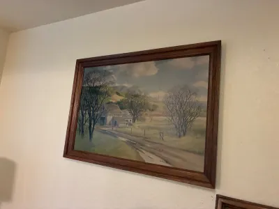 Framed landscape painting depicting a rural farmhouse, dirt road, and bare trees under a cloudy sky, presented in a dark wood frame.