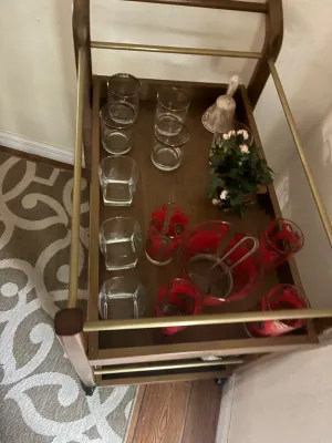 Mid-century wooden rolling bar cart featuring brass railings, loaded with various vintage glassware, including gold-rimmed rocks glasses,...