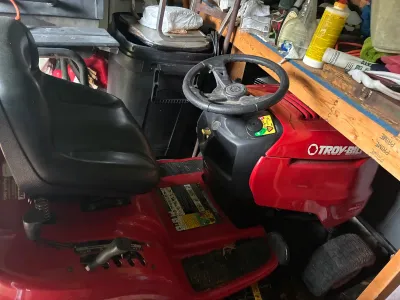 Red Troy-Bilt Bronco riding lawn tractor featuring a black high-back vinyl seat, steering wheel, and side deck controls, shown stored in...