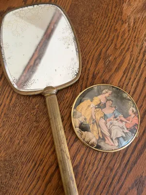 Vintage brass hand mirror and makeup compact mirror featuring a classical pastoral scene illustration.
