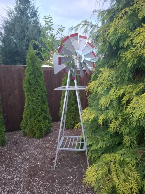 Decorative metal garden windmill with red-tipped blades, standing on a silver-painted steel frame in a landscaped yard.