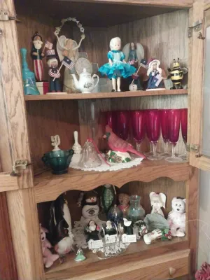 Wooden display cabinet filled with various collectibles, including patriotic figurines, a set of pink glass goblets, a cardinal figurine,...