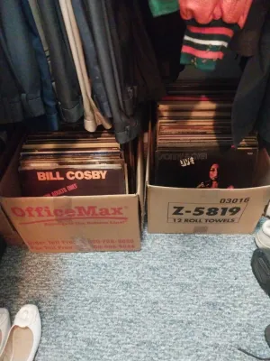Two cardboard boxes filled with vintage vinyl records, including albums by Bill Cosby and Sonny & Cher, stored on a closet floor.