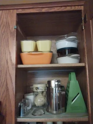 Assorted vintage kitchenware including Pyrex baking dishes, a stainless steel pitcher, a green plastic grater, and measuring cups stored...