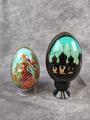 Two hand-painted Russian lacquer eggs, one depicting a woman in traditional dress carrying water buckets and the other featuring a...