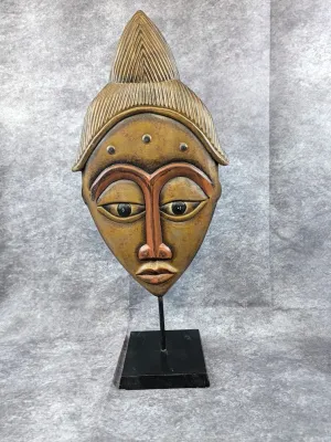 African-style carved wooden mask sculpture on a black metal display stand, featuring a pointed headdress and painted details.