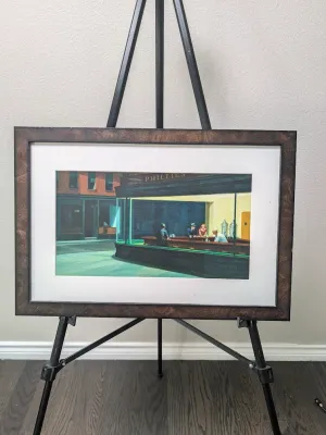 Framed print of Edward Hopper's iconic 1942 painting "Nighthawks," matted and displayed in a dark wood-grain frame on a...