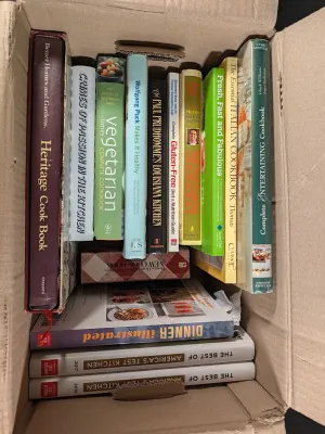 Cardboard box filled with a variety of cookbooks, including titles from America's Test Kitchen, Better Homes and Gardens, and...