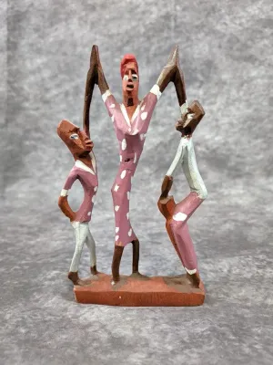 Hand-carved and painted wooden sculpture depicting three stylized figures dancing, featuring a central figure in a pink polka-dot dress...