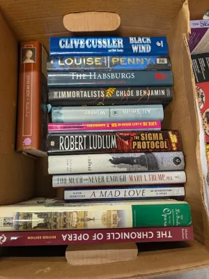 Cardboard box packed with an assortment of hardcover and paperback books, including fiction and non-fiction titles.