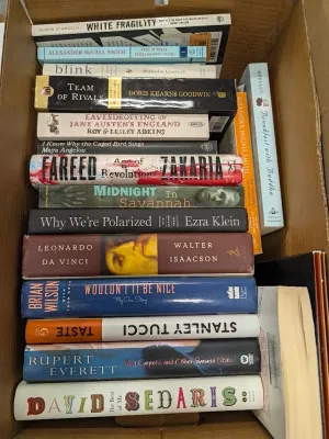 Cardboard box packed with a collection of hardcover and paperback books, including non-fiction, biographies, and contemporary literature.