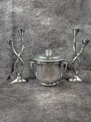 Vintage hammered aluminum ice bucket with lid and side handles, flanked by a pair of modern-style double-arm silver-tone metal candlesticks.