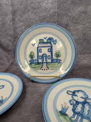 Hand-painted ceramic plates in a folk art style, featuring blue and green illustrations of a house and a sheep with blue-banded rims.