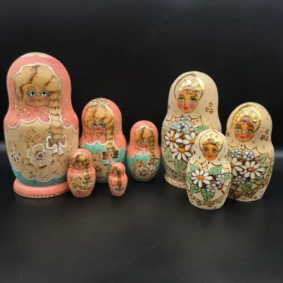 Two sets of hand-painted Russian matryoshka nesting dolls, including a pink and teal floral set and a gold-accented daisy set.