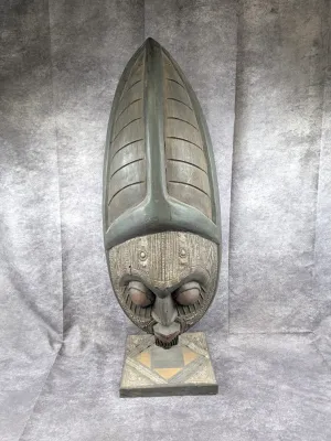 African-style carved wooden mask sculpture on a matching square pedestal, featuring intricate metal overlay details and a tall, pointed...