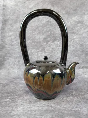 Artisan studio pottery teapot featuring a high, dramatic overhead handle and a glossy drip glaze in earth tones of brown, black, and red.