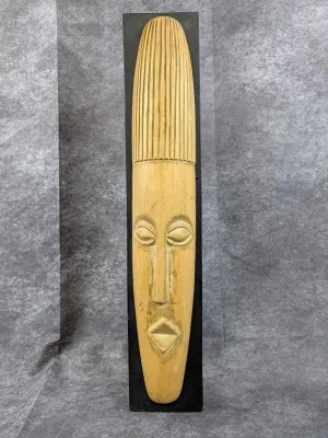Elongated African-style carved wood mask mounted on a black painted wooden display board.
