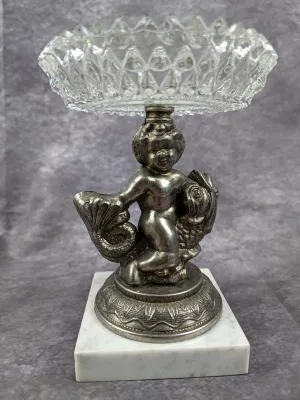 Vintage ornamental compote dish featuring a silver-toned metal cherub figure mounted on a square white marble base, topped with a...