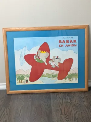 Framed "Babar en Avion" art print by Laurent de Brunhoff, featuring Babar the Elephant in a red airplane, set in a light wood...