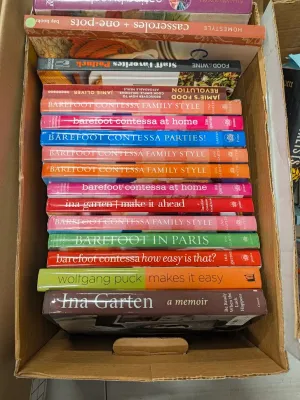 Cardboard box filled with assorted cookbooks, prominently featuring various titles by Ina Garten, along with volumes by Wolfgang Puck and...