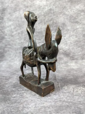 Hand-carved dark wood sculpture depicting a stylized figure riding a donkey, mounted on a rectangular wooden base.