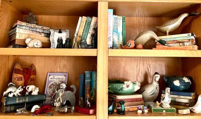 A collection of vintage books, ceramic and wooden animal figurines, and art pottery displayed on a light oak wooden bookshelf.